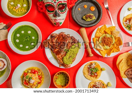 Top view photo of a red table in a Mexican restaurant with carnitas, tacos al pastor, red grasshoppers, nachos with guacamole and lots of color and a catrina mask