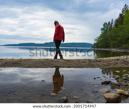 Girl wearing red jacket walking on the beach by a lake in the High Coast area, Northern Sweden. Reflections in the water, cloudy sky.