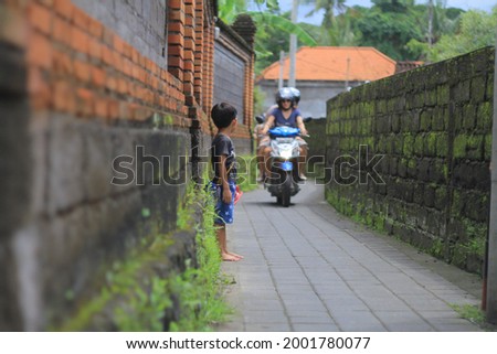 a little boy stands in a small alley in a small village in bali, waiting for a motorbike to pass