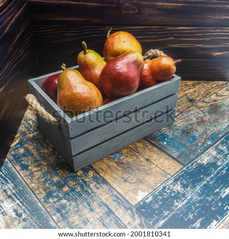 A blue wooden box with rope handles is filled with ripe pears. Red fruits with a shiny skin. Background - boards.
