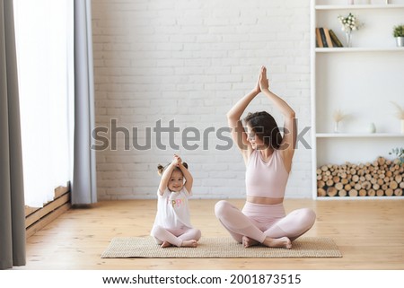 Family mother and child daughter are engaged in fitness, yoga, exercise at home. Smiling young curly woman looking at her small kids girl sitting on the carpet floor in lotus pose namaste hands up