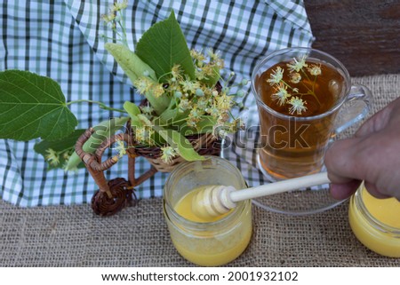 The hand takes honey with a honey stick. On jute fabric, linden flowers, jars of honey and a cup of tea with linden flowers.