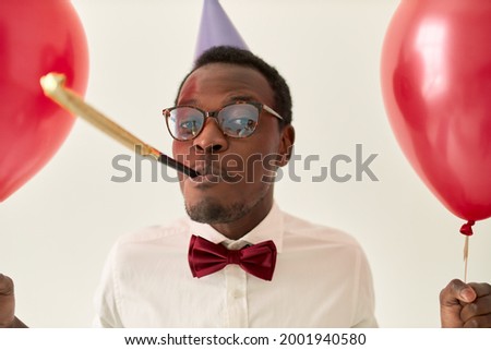 Studio image of attractive cheerful young dark skinned male wearing glasses and bow tie, whistling, celebrating birthday, holding helium balloons. Joy, happiness, festive mood and celebration concept