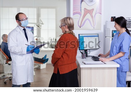 Dentist doctor with face mask explaining diagnosis to senior woman patient in stomatology waiting area hallway. Elderly man sitting on chair for teeth treatment.