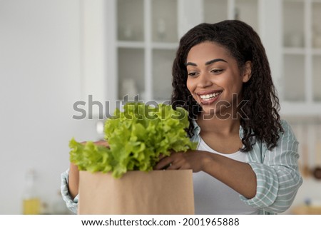 Woman sort organic food after shopping. Online buying and delivery grocery. Smiling millennial african american woman take vegetables from eco package in white kitchen interior, close up, empty space