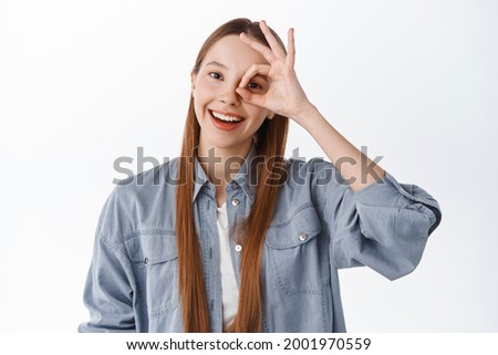 Positive teen girl, female student say okay, show OK gesture on eye and smiling, say yes, approve or recommend something cool, standing pleased against white background