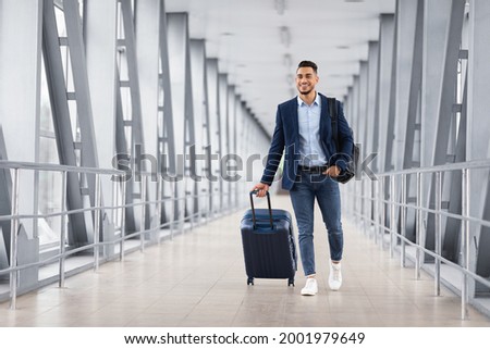 Portrait of young middle eastern man walking in airport terminal with luggage, smiling arab guy carrying suitcase while going to boarding gate, enjoying air travels and business trips, copy space Royalty-Free Stock Photo #2001979649