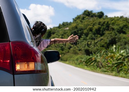 Rear side of gray car with open turn signal. Blurred of young girl sticking out of the car and showing his arms. On asphalt road with green forest and mountain for backgorund under the blue sky.
