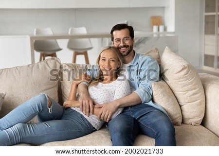 Portrait of smiling young couple hugging sitting on comfortable couch at home together, happy beautiful wife and husband in glasses looking at camera, posing for family photo in modern living room