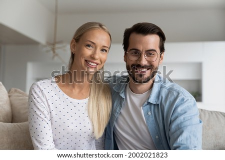Head shot portrait of smiling couple making video call together, sitting on couch at home, happy young woman and man in glasses looking at camera, chatting with friends online, using webcam