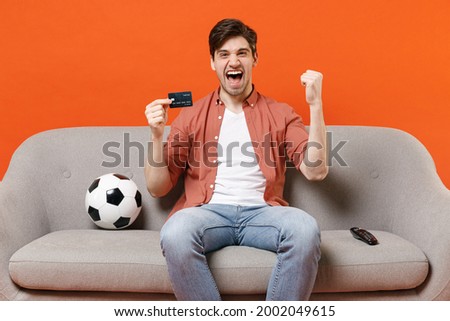 Young fun man football fan in shirt support team with soccer ball sitting on sofa home watch tv hold credit bank card do winner gesture clench fist isolated on orange background People sport concept.