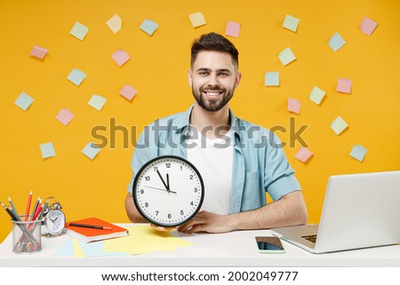Young smiling sucessful happy employee business man 20s wearing shirt sit work white office desk with pc laptop holding in hands clock look camera isolated on yellow color background studio portrait