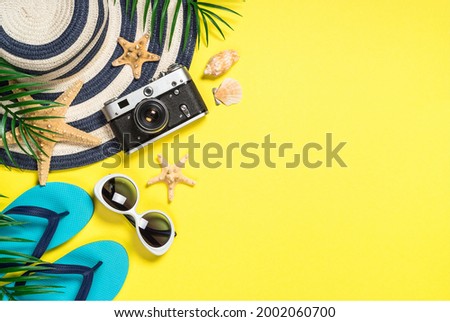 On the left side of the picture old film camera, palm leaves, and starfishes lay on hat near seashells. little far blue slippers lay near sunglasses on the yellow background color. Top views