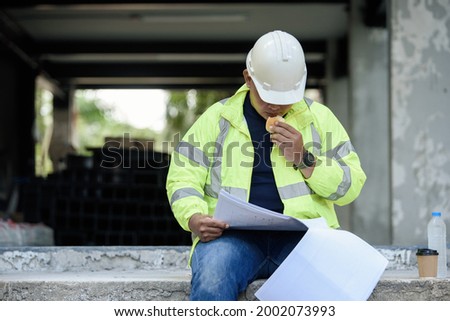 Civil Engineer sitting eats small bread in break time and reading building drawing with construction materials stock in background while hot weather at construction site during the day.