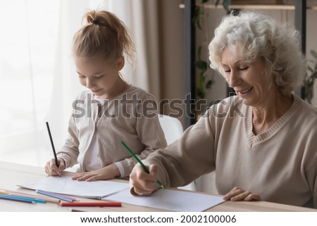 Creative activity. Happy mature grandma and preteen granddaughter friends sit at desk paint pictures in album using colored pencils. Focused little girl learn to draw imitate older female babysitter