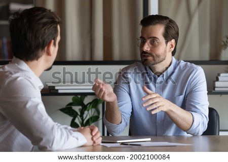 Confident advisor realtor broker manager in glasses consulting client about contract terms at meeting in office, sitting at table, two business partners negotiating, mentor teaching new employee Royalty-Free Stock Photo #2002100084