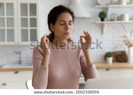 Mindful young woman breathing out with closed eyes, calming down in stressful situation, working on computer in modern kitchen. Millennial hispanic lady managing stress, practice yoga at home office. Royalty-Free Stock Photo #2002100096