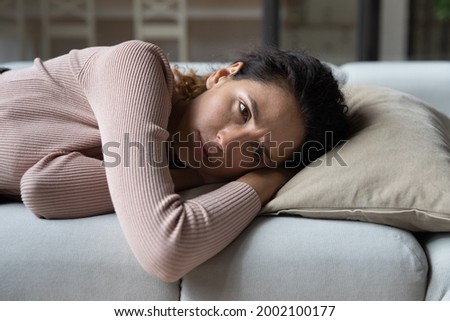 Unhappy hispanic latin caucasian woman lying on sofa, suffering from negative thoughts, getting bored, spending weekend time alone at home. Stressed millennial lady feeling unwell, depression concept. Royalty-Free Stock Photo #2002100177