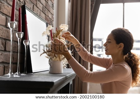 Side view beautiful young latin woman decorating fireplace with dried flowers in vase, enjoying improving or styling apartment interior, arranging indoors decor in living room, coziness concept. Royalty-Free Stock Photo #2002100180