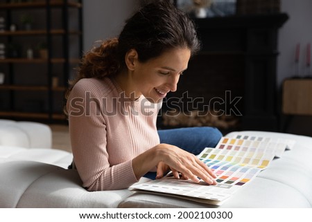 Smiling attractive millennial woman resting on cozy couch, focusing on choosing colors in swatches palette. Happy young lady planning apartment renovation, involved in creating interior design.