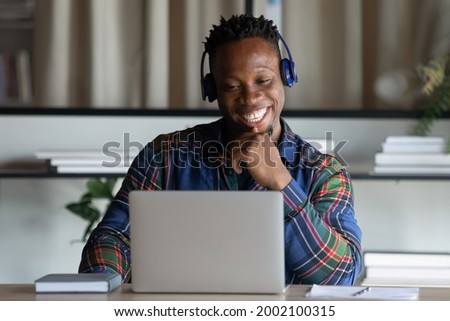 Happy African American man in wireless headphones looking at laptop screen, excited young student watching webinar, engaged in online educational course, intern studying in office, sitting at desk Royalty-Free Stock Photo #2002100315