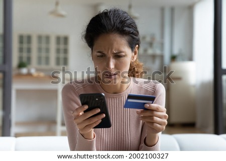 Stressed unhappy millennial woman looking at phone screen, holding bank card in hand, feeling nervous of entering wrong payment verification information, dissatisfied with service, having problems. Royalty-Free Stock Photo #2002100327