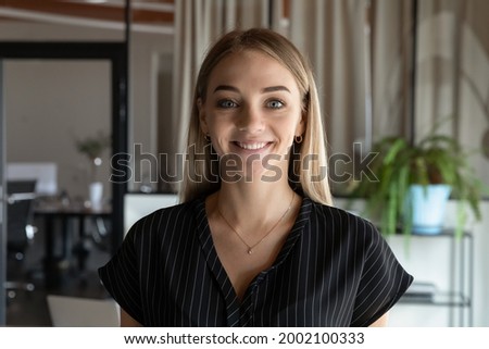 Head shot portrait close up of smiling young businesswoman intern looking at camera, standing in modern office room, successful confident employee happy female posing for corporate profile picture