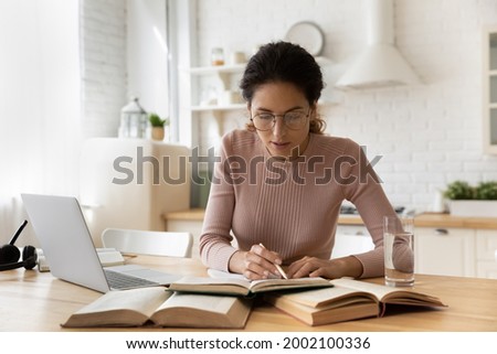 Concentrated young woman in eyewear reading carefully educational handbook, improving professional knowledge or preparing for university admission exams, writing notes in copybook, learning concept. Royalty-Free Stock Photo #2002100336