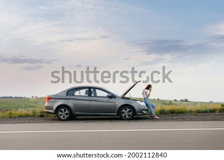 A frustrated young girl stands near a broken-down car in the middle of the highway during sunset. Breakdown and repair of the car. Waiting for help. Royalty-Free Stock Photo #2002112840