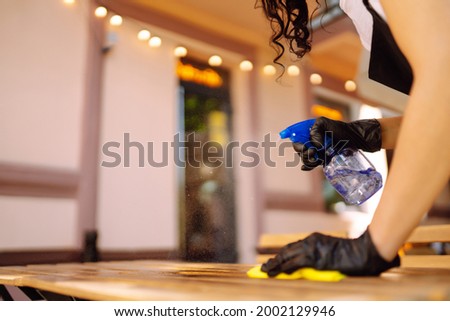 Disinfecting to prevent COVID-19. Young waitress in protective face mask and gloves cleaning table with disinfectant spray and Microfiber cloth in cafe.