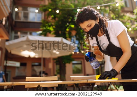 Disinfecting to prevent COVID-19. Young waitress in protective face mask and gloves cleaning table with disinfectant spray and Microfiber cloth in cafe.
