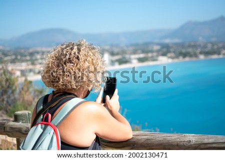 woman taking a picture of the sea with the mobile phone from a viewpoint, on a sunny day. excursion concept