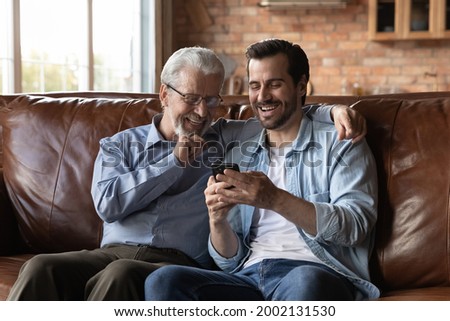 Happy senior 70s father and grown son using smartphone together, making video call, watching pictures, laughing, having fun, hugging, enjoying meeting and leisure tome at home, sitting on sofa