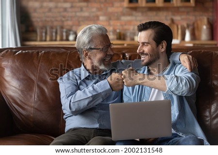 Happy excited grown son and elderly 70s father with laptop sharing good news, celebrating success, giving fist bumps. Two male generation of fans watching online sport game. Fatherhood, family leisure Royalty-Free Stock Photo #2002131548