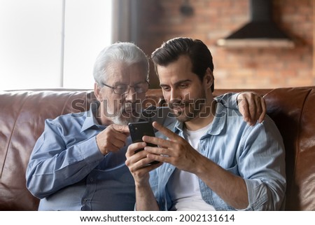 Grown son teaching elderly 70s dad to use mobile app on smartphone, showing family pictures, explaining online payment service. Two family generations men resting on couch, using cellphone together Royalty-Free Stock Photo #2002131614