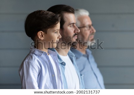 Serious thoughtful cute boy kid standing in row with young father and senior grandfather, looking forward, thinking of future. Intergenerational family portrait with three male generations Royalty-Free Stock Photo #2002131737