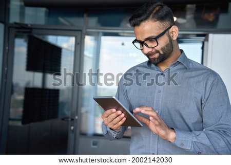Young indian marketing business manager expert in glasses standing in modern corporate office using digital tablet device searching analyzing project results. Business technologies concept. Royalty-Free Stock Photo #2002142030
