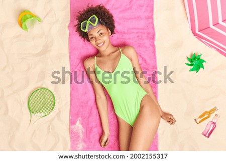 Positive relaxed curly Afro American woman in green bikini wears snorkeling mask enjoys relaxation time poses on pink towel at sandy beach. Summer time sunbathing holidays and recreation concept