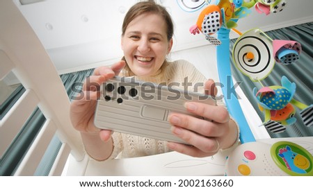 Happy smiling mother making video on smartphone of her baby lying in cradle. Concept of parenting and making images of children