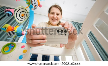 Happy smiling mother making video of her baby lying in cradle for social media. Concept of parenting and making images of children.
