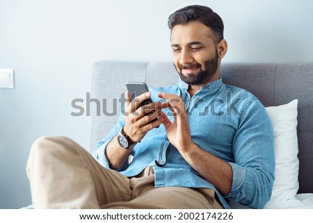 Smiling young adult indian freelance business man using mobile phone checking social media network feed or message chat sitting on bed at home. Online digital communication, rest after hard work day Royalty-Free Stock Photo #2002174226