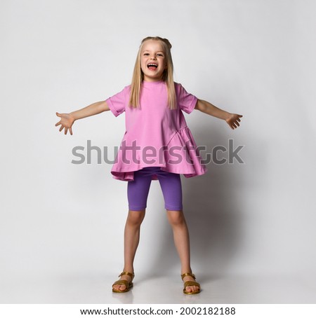 Cheerful laughing blonde 6-7 years old. a girl with a girly hairstyle in a pink sweater with a peplum and legends stands with her arms outstretched on a gray background