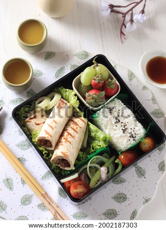 Top view of lunch box contains kebab, rice, fruit and salad. With two glass ocha tea. Selective focus.                              