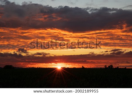 The sun sets behind shadowed fields  in Iowa.  Blue, yellow, orange and red clouds show brilliance with sun star at the horizon.  Light blue sky at the top of the image.