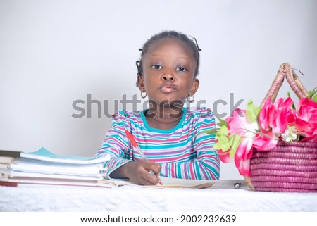 Close up education picture of an African girl child with woven hair style writing on a study table while having books and a basket of flowers beside her