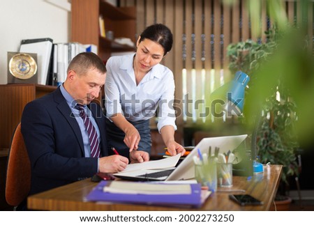 Focused adult businessman sitting at table in office, signing papers brought by female assistant