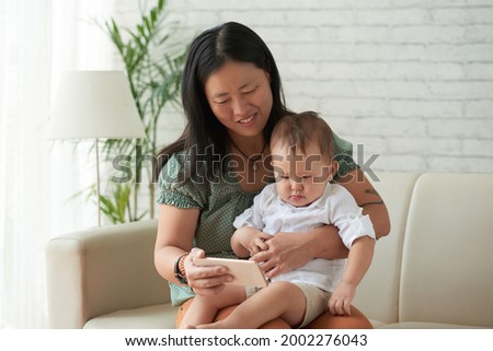 Smiling mother showing animated cartoon or educational video on smartphone to her little son when sitting on sofa at home