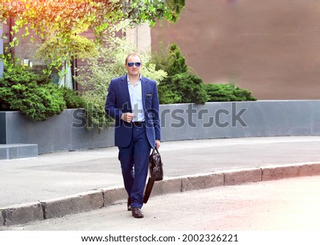 Banner with a young caucasian man in a blue business suit and sunglasses holding a suitcase walks across the road, a hurry to meet. Business, fast pace of life concept. Outside the office. 