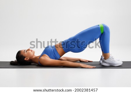 Fitness woman doing glute bridge exercise with resistance band on gray background. Athletic girl working out Royalty-Free Stock Photo #2002381457