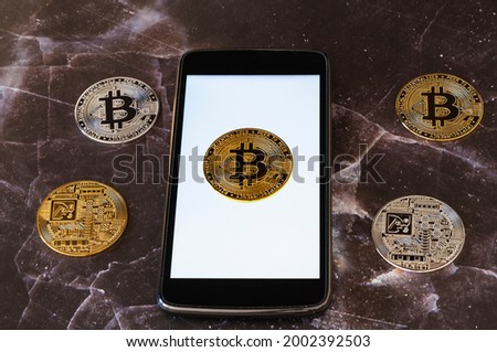 On a dark marble surface several physical bitcoin cryptocurrencies. One of the coins is on top of the white lit screen of a cell phone.